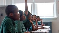 Six high school students, all in green shirts, are sitting in a row at individual desks looking towards the front of the class. One of them has their hand raised.