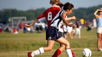 Two teen girls are fighting for a ball on the soccer field.