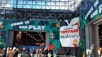 People are outside, walking towards a building that says "Come and Play," and "Welcome to Toy Fair NY 16."