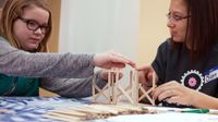 Two girls are sitting, building a house with popsicle sticks on a table with a white and blue checkered cover.