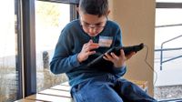 A young boy in a blue sweater and jeans with glasses is sitting on a bench indoors beside a window, holding a tablet and looking at a sheet of paper on the bench.