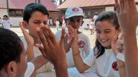 In the background, a lot of young students are playing outside on cement, closed in by their surrounding classrooms. At the forefront, five students are sitting around a small rectangular table with their elbows on the table and their hands raised, smilin