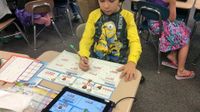 A young boy is sitting at his desk in class in a yellow, long-sleeve Minion shirt. He's wearing headphones connected to a tablet, and is doing math problems on paper that matches what he sees on the tablet.