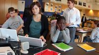 Three students are sitting at a long, black table in a large classroom. Two are on laptops, and one has her face in her hands with her elbows on the table. More students are sitting behind them at desks and a teacher is standing behind them.
