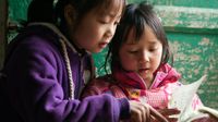 Two young girls, one in a purple sweatshirt and one in a pink sweatshirt, are sitting outside against a green, wooden door, reading together.