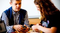 Two teenagers are sitting at a table with a laptop, one is holding a cell phone charger and the other is holding a cell phone, connecting it to a white wire.