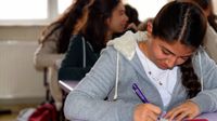 A closeup of a high school girl sitting at a purple desk. She's writing, looking down at her paper. Other students are sitting behind her, slightly blurred.