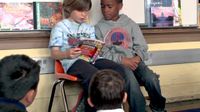 Two young students are sitting in chairs against the wall with a whiteboard behind them. They're reading a book to the rest of the class who are sitting on the carpeted floor in front of them.