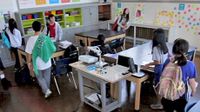 Seven students are walking into a classroom, setting their stuff down at tables.