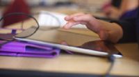 With a blurred background, a student's hand is highlighted hovering over a tablet.