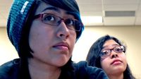 A closeup of two female teens from the shoulder up sitting, looking towards the front of the class.