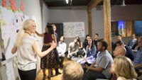 A group of teachers are sitting in a large room, and three teachers are presenting next to a post-it and paper covered wall.
