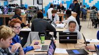 A classroom filled with high school students sitting at large rectangular tables, working together on laptops.
