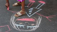 A girl in golden sandals and a colorful polka dot dress is standing on cement, bending over, holding pink chalk, drawing a smiling girl.