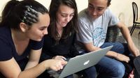 Three high-school teens are sitting next to each other on a couch, two girls on the left and a boy on the right. The girl in the middle has a laptop on her lap that they're all looking at.