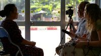 Three adults are sitting across from an old woman in a classroom, filming her. One of them is holding a camera on a tripod, and another is holding a long microphone.