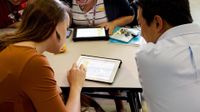 Four teachers are sitting around a table, looking at spreadsheets together on two tablets