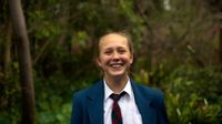A young, teenage girl in a white collared shirt, blue and red checkered tie, and a blue jacket is standing, smiling, with trees as her backdrop.
