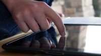 A closeup of a person's hand pressing their index finger against a tablet on a wooden table. In the upper right corner of the photo, there is a blurred corner of brick house and a tall green bush.