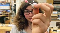 A curly-haired teenage girl is standing in a classroom holding a metal circle that's connected to two miniature red light bulbs. One of them is lit.