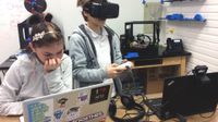 A young girl is sitting at a classroom table working on her laptop. A young boy is standing next to her with high-tech eye goggles on, a video game controller in his hands, and he's looking at a laptop with a video camera connected on the top.