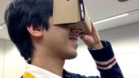 A teenage boy is holding a Google Cardboard viewer up to his eyes, a cardboard box with his smart phone inside of it, looking into it as if it were binoculars or a camera.