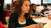 A young girl is sitting in class, looking straight ahead with a playing card in her hand and a green sheet of paper on her desk. Other students in the classroom are sitting with green sheets of paper and playing cards about.