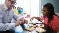 A female student and a male teacher are sitting at a raised table together putting wires together to turn on small light bulbs connected to square pieces of wood.