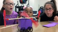 Three upper elementary girls are squatting next to a table, blowing on the wings of a toy car they've made, propelling it to move.