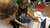 Three young teens are standing, hovering over a long, brown desk with folders, a red bin filled with notebooks, a pair of scissors, and opened folders with papers strewn across it. The girl is leaning towards a laptop, typing.