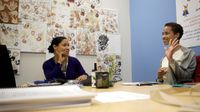 Two female teachers are sitting at a large table in a classroom. They're smiling and talking.