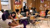 A group of students are sitting at their desks in class, listening to their teacher speak.