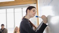 A high school female is holding a notebook in one hand and writing on a whiteboard at the front of the class with another hand. Two other students are sitting at a table nearby her.