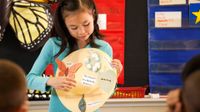 A young girl is standing at the front of a classroom holding a collage that represents the life cycle of a butterfly. She's presenting on it to her class.