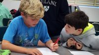 Amidst other students, two young boys are sitting at their classroom table, working on an assignment together.