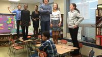 Six preteen students are standing on their classroom desks, smiling, and one student is sitting.