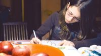 A young girl with glasses is sitting at her kitchen table writing. There's a bowl of apples on the table, next to an opened text book, next to an opened binder with lined paper.