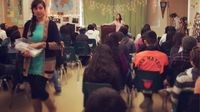 A high school classroom is decorated with white strands of lights, and a green sheet backdrop partially covering the front of the classroom. The classroom is filled with students sitting in blue chairs. One teacher is standing at a podium, addressing the