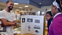A teacher is smiling at two young students, standing in front of a cardboard display on a desk.