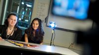 Two young girls are sitting at a table in a classroom being filmed with a light behind them.