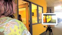 A tripod is standing in the middle of a school hallway with an iPad on it. A student is standing behind it, looking off to the side. Another student is standing between a doorway, looking into the hallway.