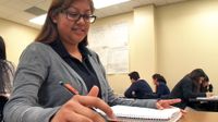 A closeup of a teenage girl sitting at her desk, smiling. One hand is resting on an opened notebook, and her other hand is holding a pencil above a sheet of paper she's writing on. Other students are sitting throughout the classroom.