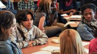 A group of ten high school students are in a library sitting at tables and talking to each other.