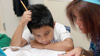 A young boy is sitting at his desk beside his teacher. His left forearm is on his desk, his head resting against it. His right elbow is resting on his desk with his hand against his head. He's looking at his open notebook, working with his teacher.