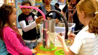 Three elementary-aged girls and two female teachers are standing around three rectangular desks pushed together with a student-made contraption standing on the center desk. The contraption is a large, circular, aluminum-looking tube standing about two to