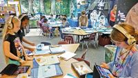 Tablets and lamented information sheets next to boxes filled with marine life span across nine classroom tables. Seven students are sitting at one table, two students are standing together, and two others wearing headphones are working individually.