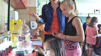 Teacher standing talking with a student as they watch a girl pouring liquid into cups at a counter