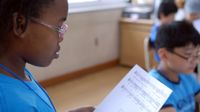 A closeup, side profile of a young girl with glasses standing in a classroom, reading a music sheet.