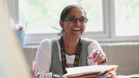 A female teacher is sitting at a desk, looking up and smiling. She's holding a pen in one hand, and stacks of paper and folders are in front of her. She's wearing glasses, big, pearl-like earrings, a turquoise necklace, and a lanyard.