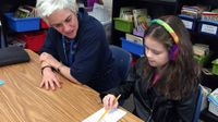 An young girl with rainbow-knitted headphones is sitting at a table with her female teacher. They're both looking at a long, thin sheet of paper, and the young girl is holding the tip of the pencil against the paper, pointing at something.